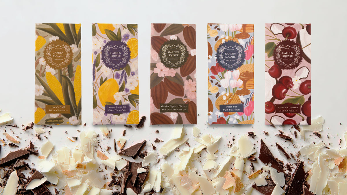 Five Garden Square Chocolatier bars—Iowa's Gold, Lemon Lavender, Classic, Dutch Bar, and Brandied Cherries—stand above a pile of white and dark chocolate shavings.