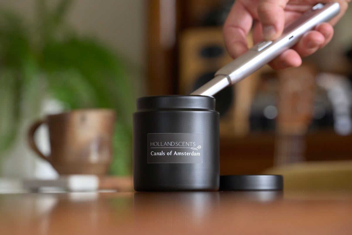 Holland Scents Canals of Amsterdam soy candle being lit on a wooden table with a coffee mug and plant, Pella, Iowa